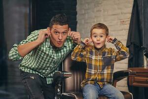 stylish little kid sitting on chair at barbershop with his young father on background photo