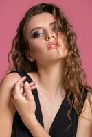 Beautiful fashionable girl with long curly hair in a black dress in the studio on a pink background. photo