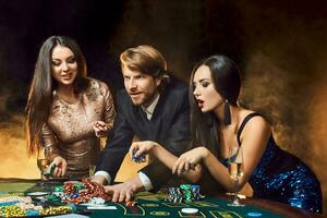 Two beautiful women and young man play on poker table in casino, focus on man and brunette photo