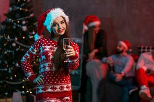 Portrait of a young woman with a glass of champagne at home on the foreground. Beautiful brunette in a Santa hat, red costume with deers photo