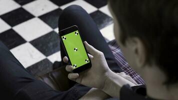 Over the shoulder view of a man using smart phone with green screen in the room with chessboard floor. Stock footage. Male holding his device with chromakey screen and looking at it. photo