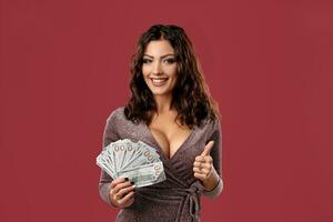 Brunette girl in shiny dress posing holding a fan of hundred dollar bills in her hand, standing on pink studio background. Casino, poker. Close-up. photo