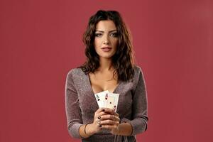 Brunette girl in a shiny brown dress posing holding two playing cards in her hands, standing against pink studio background. Casino, poker. Close-up. photo