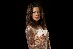 Brunette girl wearing shiny dress posing holding two playing cards in her hand standing against black studio background. Casino, poker. Close-up. photo