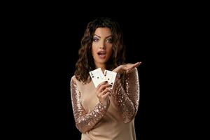 Brunette girl wearing shiny dress posing holding two playing cards in her hand standing against black studio background. Casino, poker. Close-up. photo