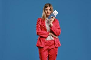 Blonde lady in white blouse and red pantsuit. She is holding passport and ticket while posing on blue background. Travelling concept. Close-up photo