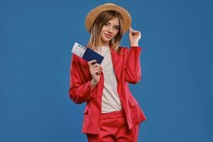 Blonde model in straw hat, white blouse, red pantsuit. She is holding passport and ticket, posing on blue background. Travelling concept. Close-up photo