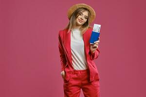 Blonde model in straw hat, white blouse and red pantsuit. She is holding passport and ticket while posing against pink studio background. Close-up photo