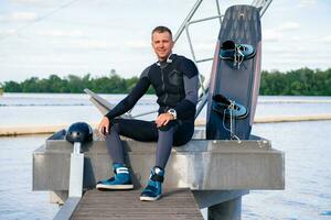 Smiling confident wakeboarder resting after training, sitting on pier with wakeboard photo