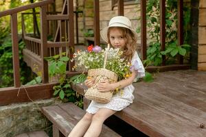 linda pequeño niña sentado en peldaño de país casa con cesta de flores silvestres foto