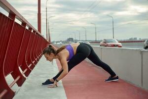 niña atleta extensión en el Mañana afuera, en el ciudad, en moderno puente cruce río. activo sano estilo de vida. deporte y recreación concepto. foto