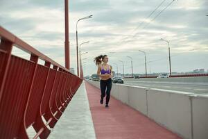 Girl athlete is jogging in the morning outside, in the city, on modern bridge crossing river. Active healthy lifestyle. Sport and recreation concept. photo