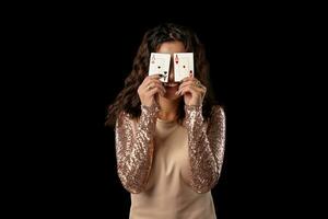 Brunette girl wearing shiny dress posing holding two playing cards in her hands standing against black studio background. Casino, poker. Close-up. photo