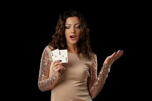 Brunette girl wearing shiny dress posing holding two playing cards in her hand standing against black studio background. Casino, poker. Close-up. photo