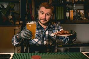simpático barbado barman ofrecimiento vaso de cerveza con meriendas en pub foto