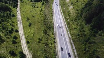 Aerial top view of summer green trees, river, roads in forest background. Aerial view of crooked path of road on the mountain. Clip. Aerial view of cars driving on country road in forest. The aerial photo