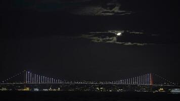 Beautiful illuminated bridge above the river on the city lights background. Action. Night cloudy sky and the moon. photo