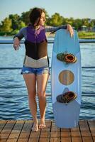 Girl in a gray turtleneck, special sports vest and blue denim shorts is looking away while posing with her wakeboard on a pier. Full length shot. photo