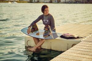 Brunette girl in a gray turtleneck and blue denim shorts is posing with her wakeboard on a pier of the coastal zone. photo