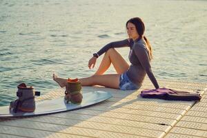 Brunette girl in a gray turtleneck and blue denim shorts is posing with her wakeboard on a pier of the coastal zone. photo