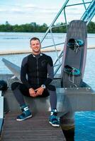 Cheerful wakeboarder relaxing after training sitting on pier with board photo