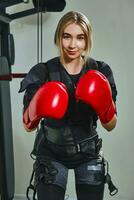 hermosa rubia mujer vistiendo en sensores y cinturones, sujeta con velcro es practicando ems aptitud en un gimnasio utilizando un rojo boxeo guantes. foto