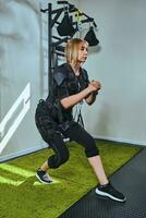 Beautiful blonde woman wearing in sensors and belts, fastens with velcro is practicing EMS fitness in a gym, doing squat. Modern kind of sport. photo