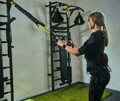 Beautiful blonde woman wearing in sensors and belts, fastens with velcro is practicing EMS fitness in a gym performing traction exercises. photo