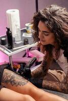 hembra tatuaje artista utilizando máquina a crear flor diseño en cliente muslo foto