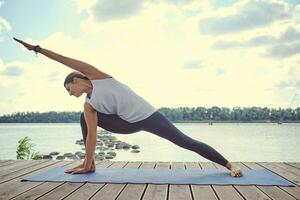 Woman in white undershirt and black leggings is practicing yoga performing yoga-asanas on a mat outdoors, near a riverside. photo