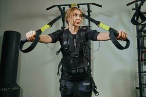 Beautiful blonde woman wearing in sensors and belts, fastens with velcro is practicing EMS fitness in a gym performing traction exercises. photo