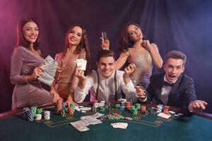 Friends playing poker at casino, at table with stacks of chips, money, cards on it. Celebrating win, smiling. Black, smoke background. Close-up. photo