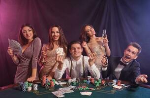 Friends playing poker at casino, at table with stacks of chips, money, cards on it. Celebrating win, smiling. Black, smoke background. Close-up. photo