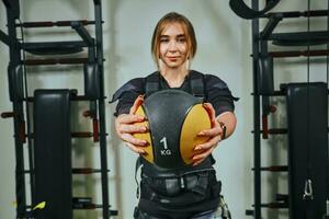 Beautiful blonde woman wearing in sensors and belts, fastens with velcro is practicing EMS fitness in a gym, building up muscles with a weighted ball. Modern kind of sport. photo