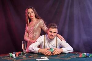 Man playing poker at casino, sitting at table with stacks of chips and cards on it. Celebrating win with woman. Black, smoke background. Close-up. photo