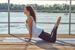 Woman in white undershirt and black leggings is practicing yoga performing yoga-asanas on a mat outdoors, near a riverside. photo