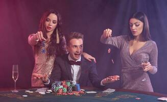 Man playing poker at casino sitting at table with stacks of chips, money, cards. Celebrating win with two women. Black, smoke background. Close-up. photo