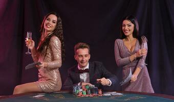 Man playing poker at casino sitting at table with stacks of chips, money, cards. Celebrating win with two women. Black background. Close-up. photo