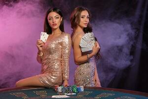 Two women showing cards and money, posing at playing table in casino. Black, smoke background with colorful backlights. Gambling, poker. Close-up. photo