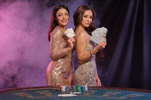 Two women showing cards and money, posing at playing table in casino. Black, smoke background with colorful backlights. Gambling, poker. Close-up. photo