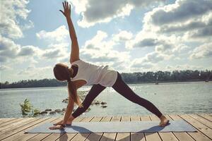 Woman in white undershirt and black leggings is practicing yoga performing yoga-asanas on a mat outdoors, near a riverside. photo