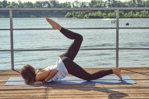 Woman in white undershirt and black leggings is practicing yoga performing yoga-asanas on a mat outdoors, near a riverside. photo