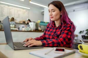 Young woman with pink hair with laptop computer sitting in cafe, intelligent female student working on net-book. photo