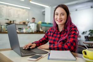 Young woman with pink hair with laptop computer sitting in cafe, intelligent female student working on net-book. photo