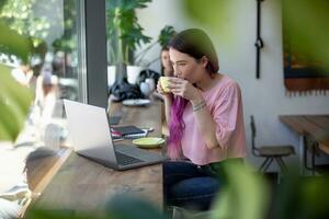 Side view of young businesswoman sitting at table in coffee shop. On table cup of coffee and laptop. In background white wall and window. photo