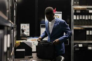 African american detective preparing his backpack, leaving job late at night in arhive room. Police officer working overtime at criminal case, analyzing crime scene evidence. Criminology concept photo