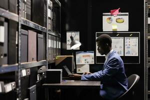 African american police officer analyzing investigation files, working overtime at confidential case in arhive room. Private detective checking victim evidence, planning strategy to catch suspect. photo