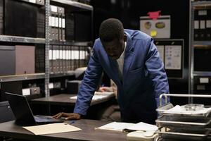 African american officer analyzing criminal case, checking crime scene evidence in arhive room. Private detective reading criminology report, searching for clues. Law enforcement concept photo