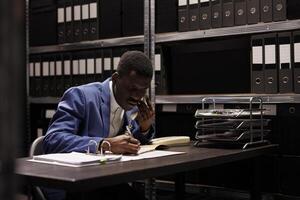 African american bookkeeper having remote discussion with manager using landline phone, discussing bookkeeping report in storage room. Businessman working overtime with administrative files photo