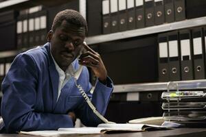 Police officer explaining confidential case to remote inspector using landline phone, working overtime in arhive room. Private investigator checking crime scene evidence. Investigation concept photo